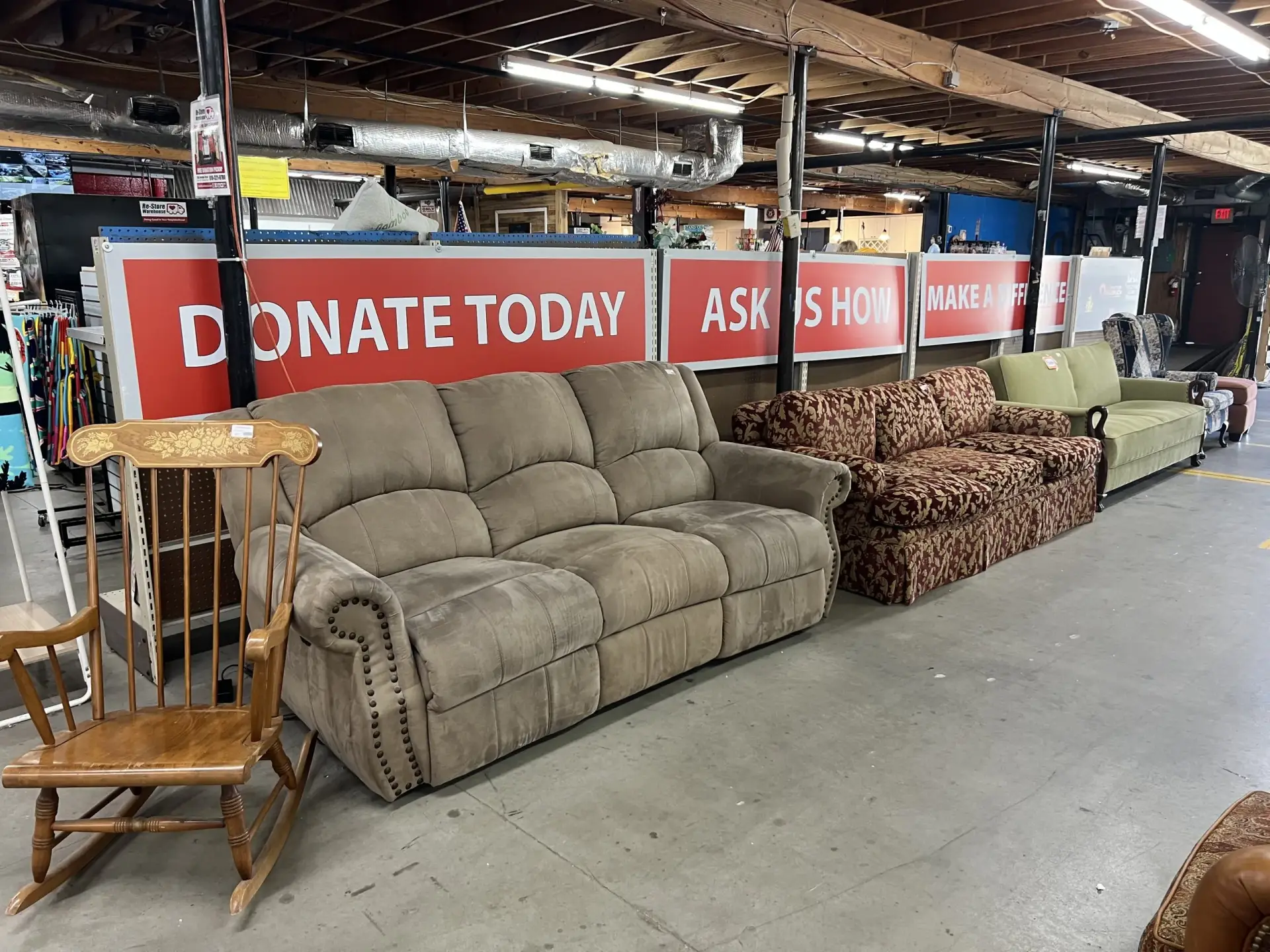 A showroom aisle featuring a wooden rocking chair and a row of sofas—including a brown microfiber couch and a patterned red sofa—lined up in front of large red and white signs that read "DONATE TODAY," "ASK US HOW," and "MAKE A DIFFERENCE."