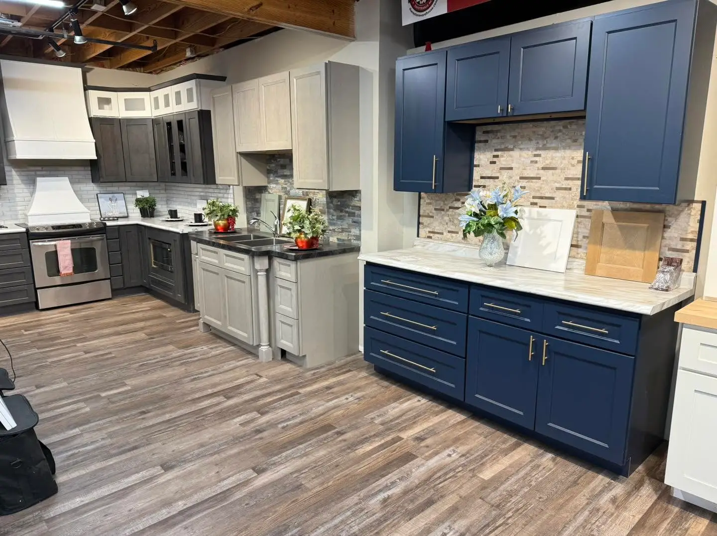 A showroom display featuring multiple styles of kitchen cabinetry installations, including a modern navy blue cabinet unit with gold hardware, a light grey island with a double sink, and a dark charcoal corner kitchen setup with a stainless steel range, all on a wood-look plank floor.