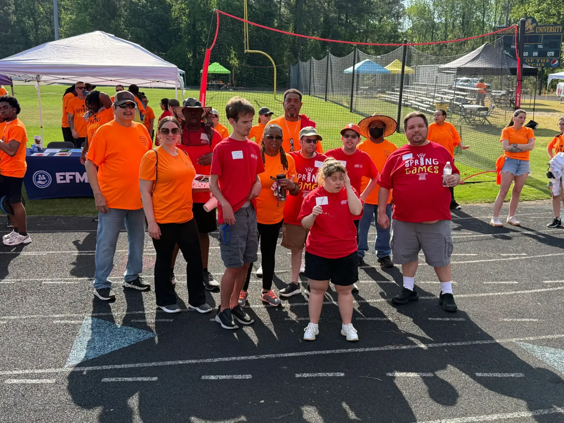 A group of participants and volunteers wearing bright orange and red "Spring Games" t-shirts standing together on an outdoor running track at Methodist University during a community sports event.