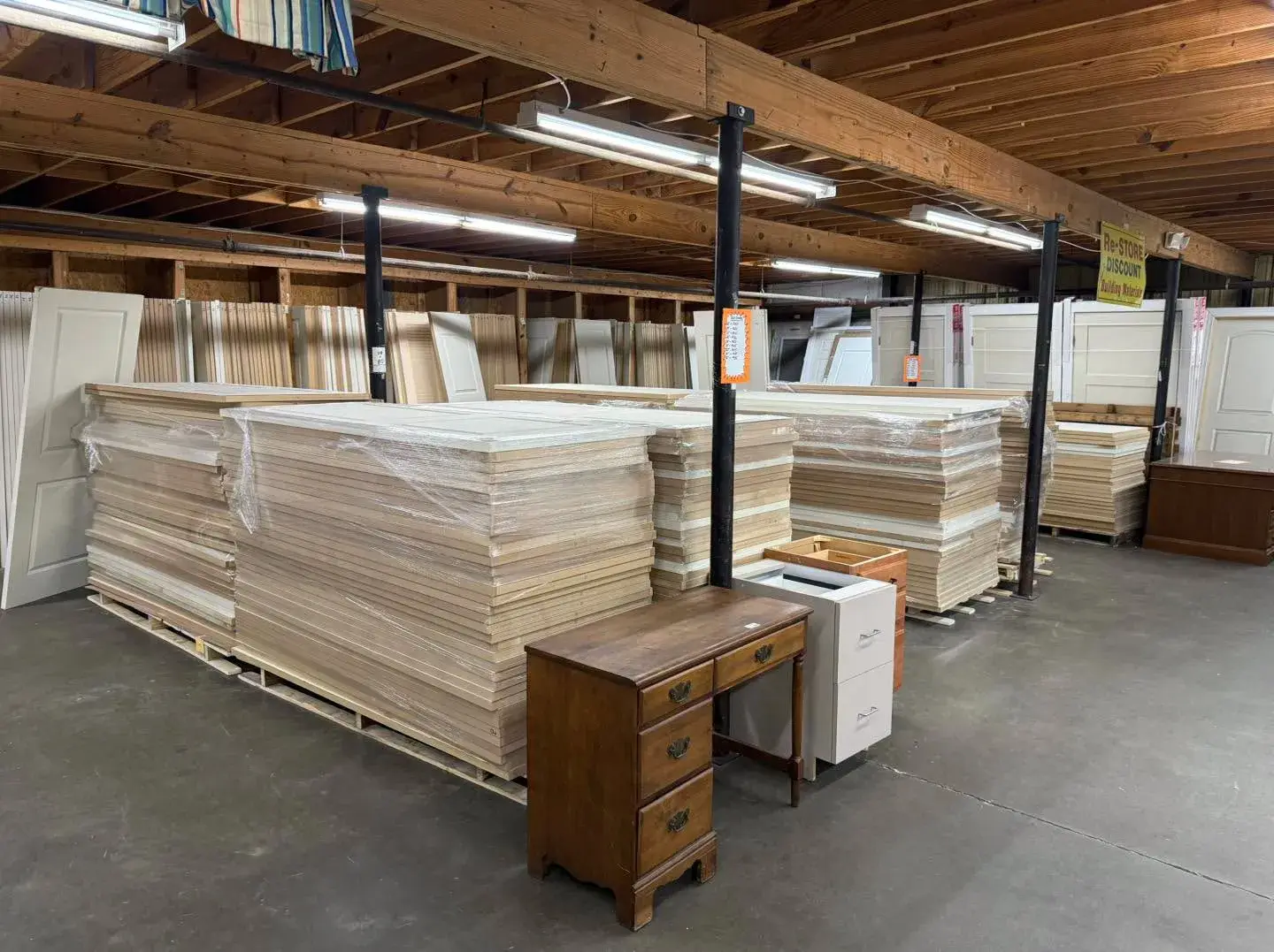 Large stacks of shrink-wrapped wooden doors and building materials displayed inside Re-Store Warehouse alongside a wooden desk