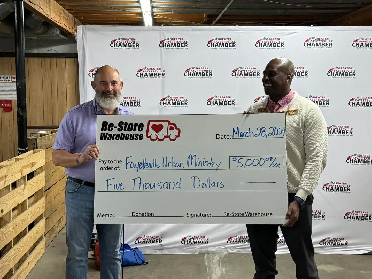 Two men standing in front of a Greater Fayetteville Chamber backdrop holding a large novelty check for $5,000 from The Re-Store Warehouse to Fayetteville Urban Ministry.