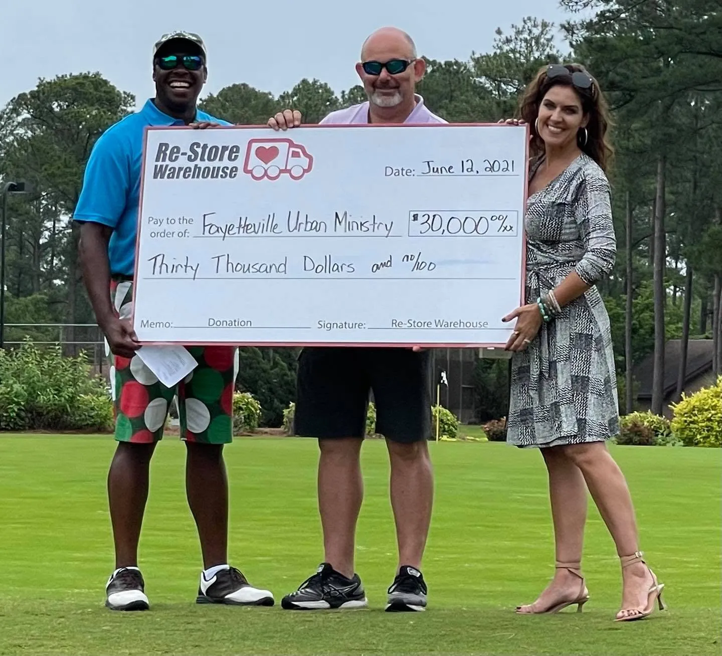 Three representatives standing on a golf course green holding a large $30,000 donation check from Re-Store Warehouse to Fayetteville Urban Ministry.