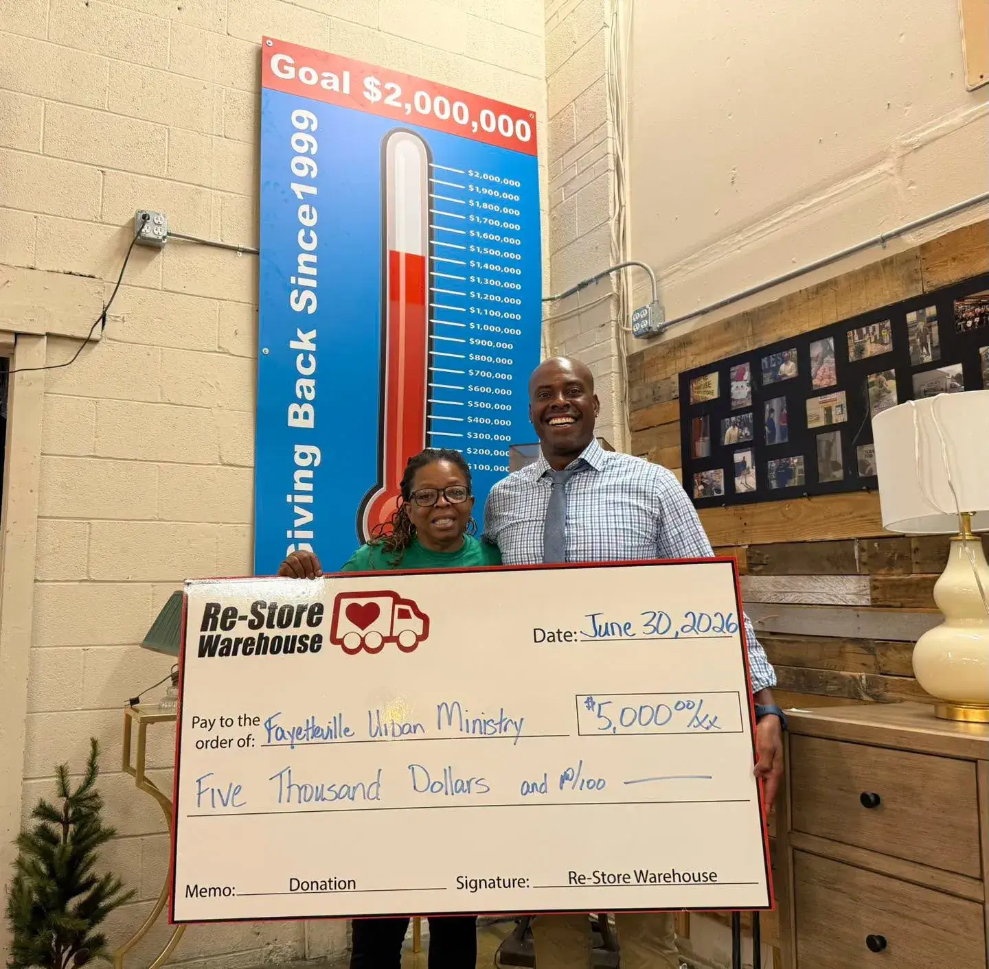 Two representatives holding a large $5,000 donation check from Re-Store Warehouse to Fayetteville Urban Ministry in front of a fundraising thermometer banner.
