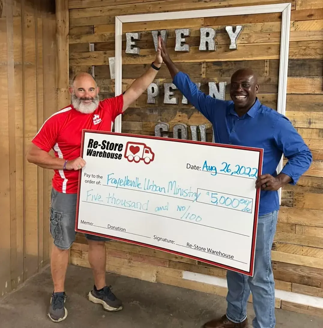 Two men high-fiving while holding a large $5,000 donation check from Re-Store Warehouse to Fayetteville Urban Ministry.