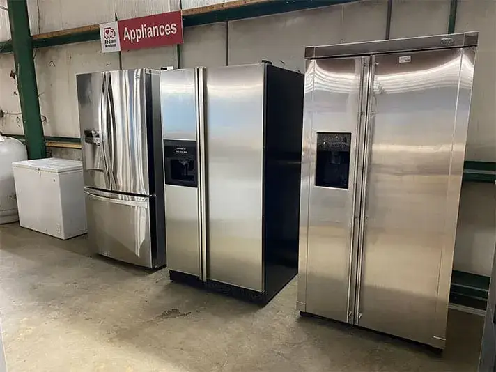 A selection of stainless steel refrigerators, including a French door model and side-by-side units, lined up on a concrete floor in an appliance section.