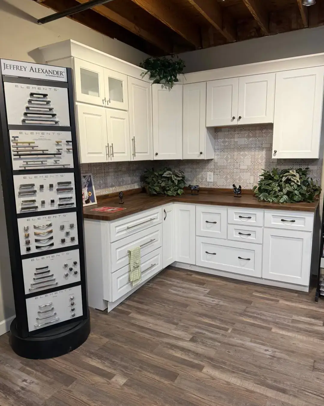 White shaker-style kitchen cabinetry with butcher block countertops next to a Jeffrey Alexander cabinet hardware display stand.