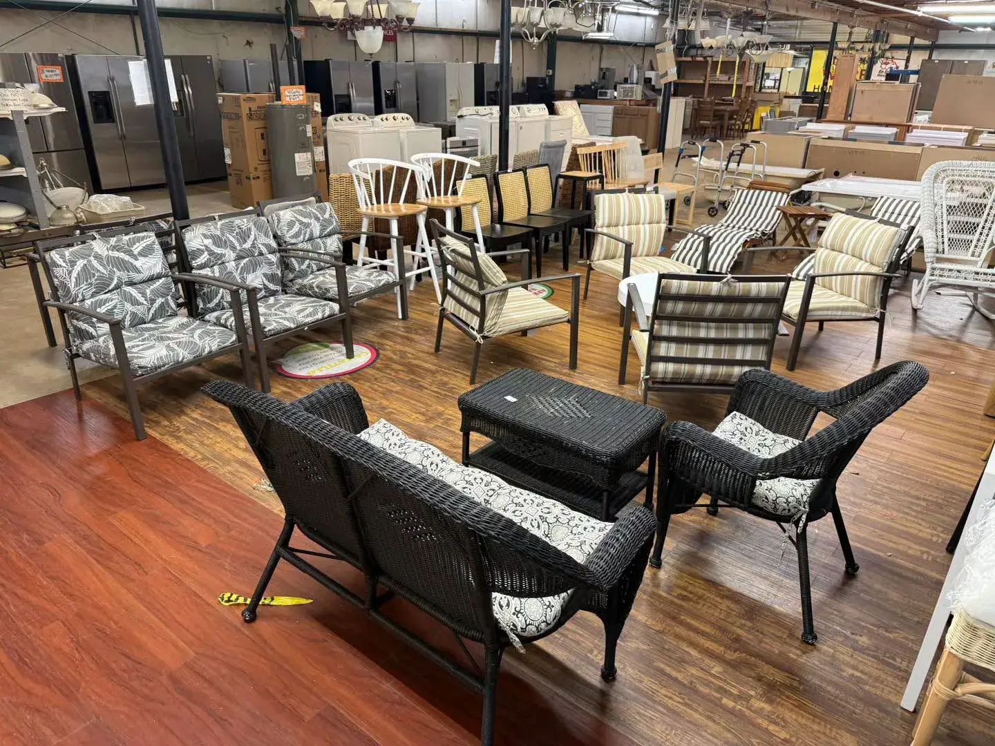 Outdoor patio sets including a black wicker loveseat set, palm leaf cushioned armchairs, and striped lounge chairs displayed on wooden flooring inside The Re-Store Warehouse.